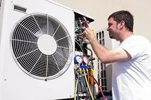 HVAC technician repairs an air conditioning unit outside. He uses tools on the open panel with a gauge connected, and smiles.