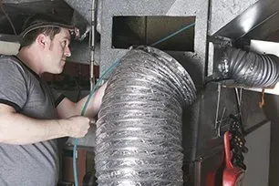 Man connecting flexible ductwork to metal duct in a basement; work light on his head.