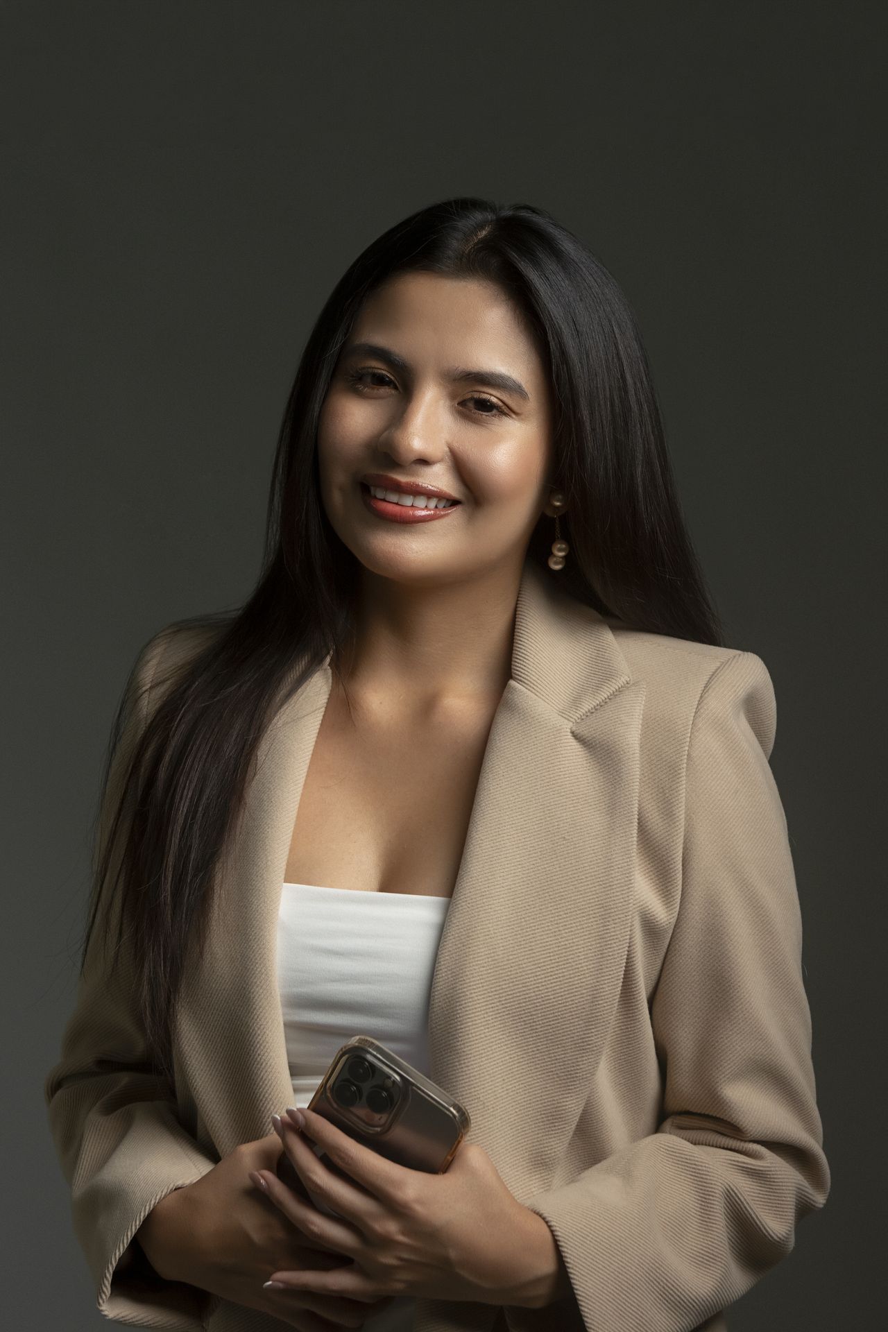 Mujer con blazer beige sosteniendo un teléfono, sonriendo contra un fondo de estudio oscuro.