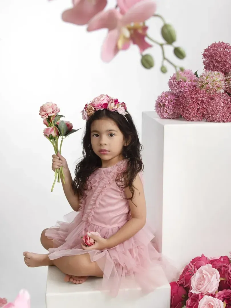 Niña pequeña con un vestido rosa sentada entre flores rosas, sosteniendo un ramo en un ambiente de estudio tranquilo.