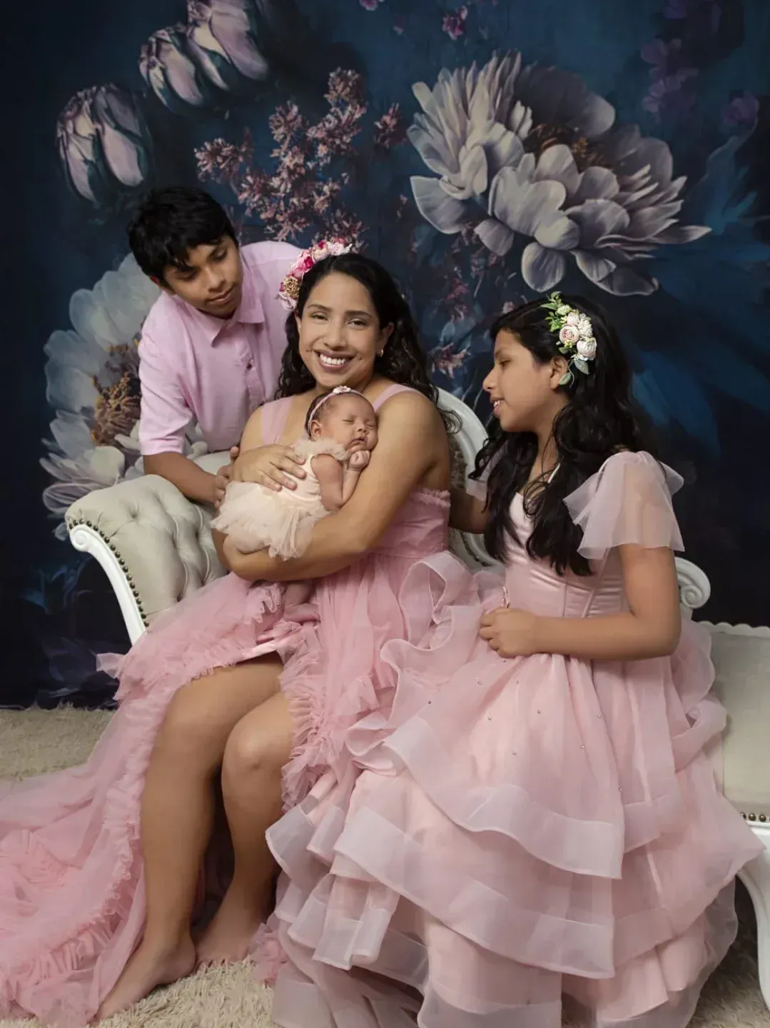 Retrato familiar en una silla rosa, con la madre sosteniendo a un bebé y dos niñas con vestidos rosas sobre un fondo floral.