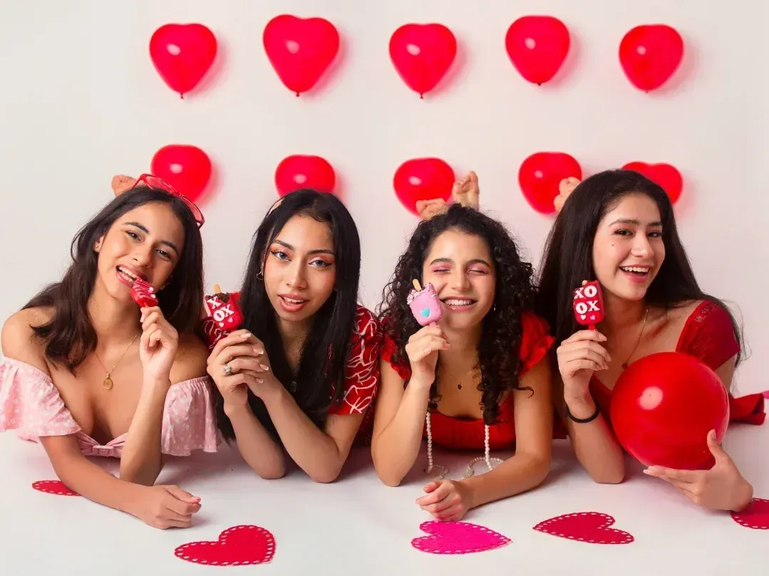 Cuatro personas con globos rojos en forma de corazón y caramelos, sonriendo en una mesa decorada para el Día de San Valentín.