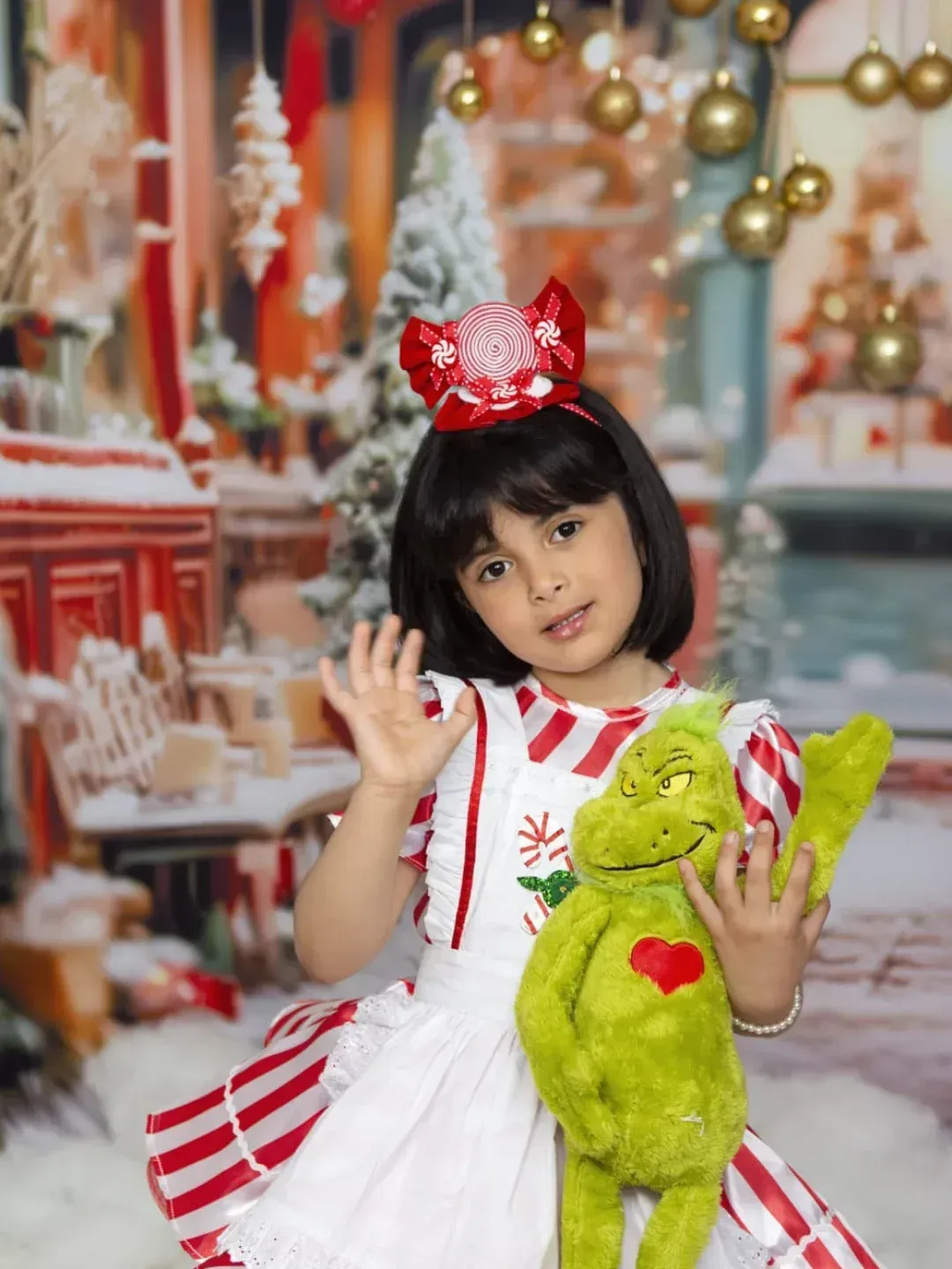 Una niña con un lazo rojo y un vestido blanco sostiene un osito de peluche verde en una escena festiva navideña.