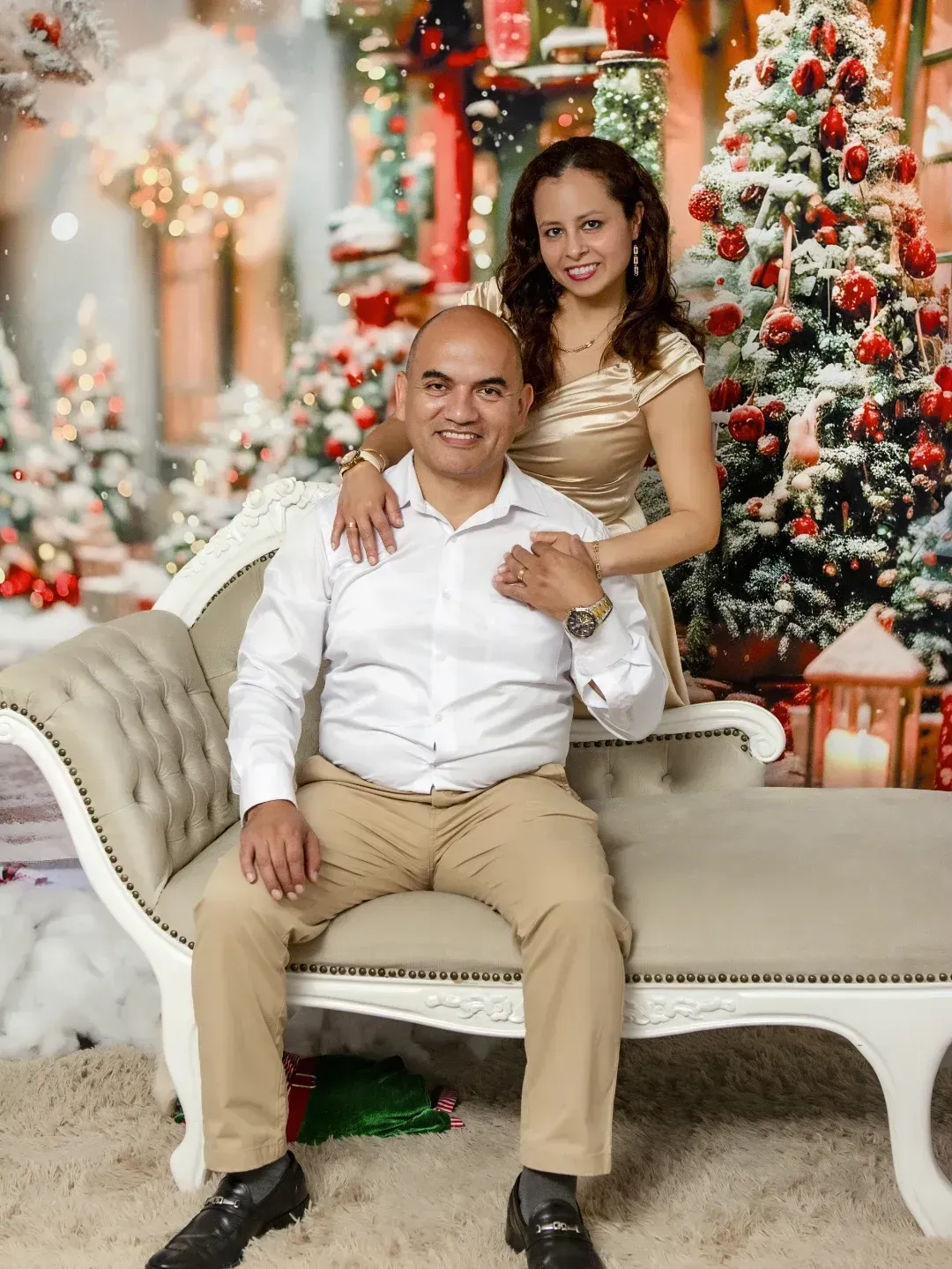 Pareja posando en un sofá color crema en un ambiente festivo navideño con árboles de Navidad decorados y luces cálidas.