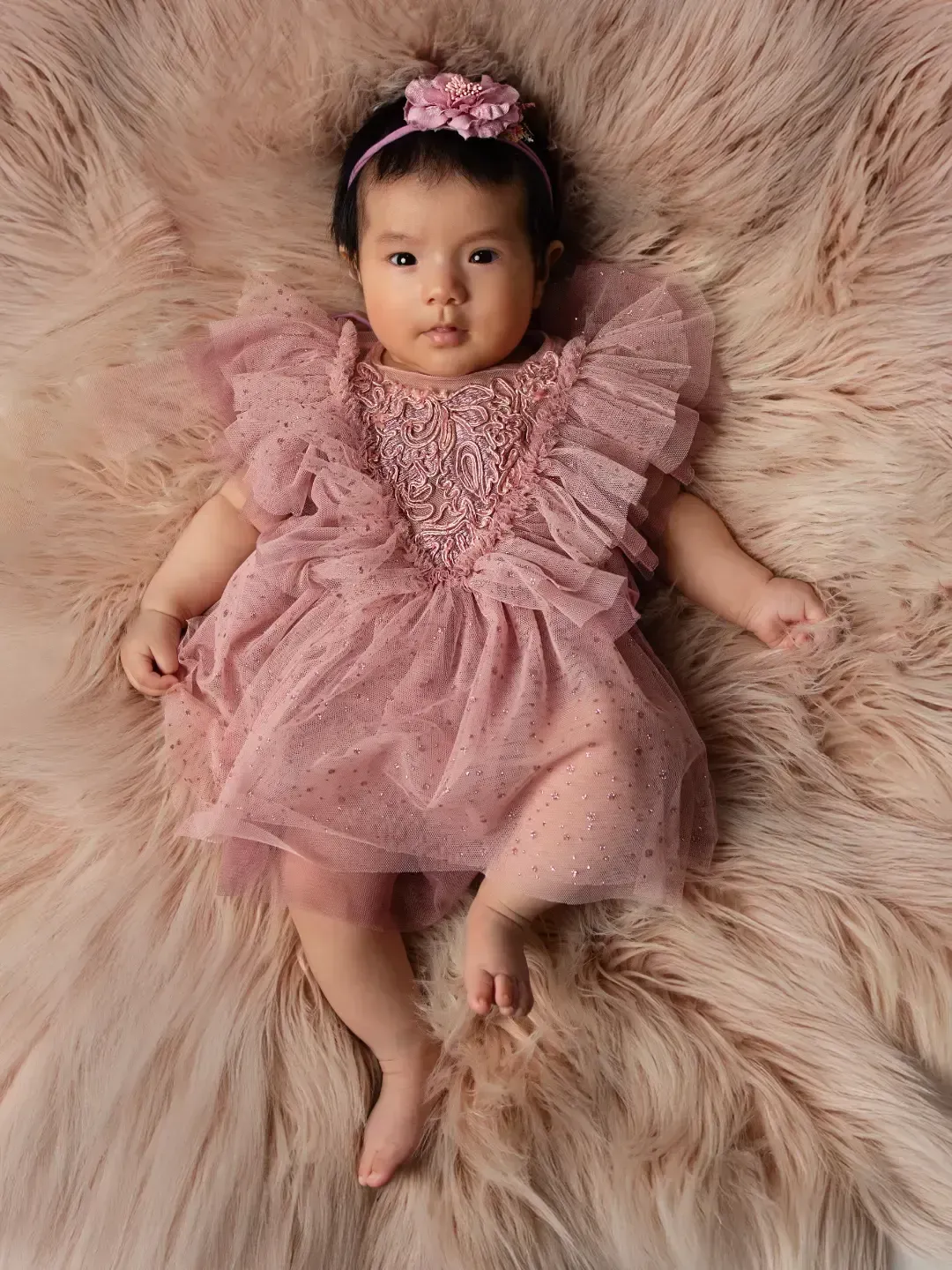 Bebé tumbada sobre una alfombra mullida de color beige, con un vestido rosa y una diadema floral.
