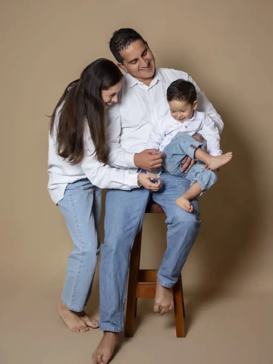 Tres personas con camisetas blancas y vaqueros azules están sentadas juntas en un taburete de madera, sonriendo contra un fondo beige.