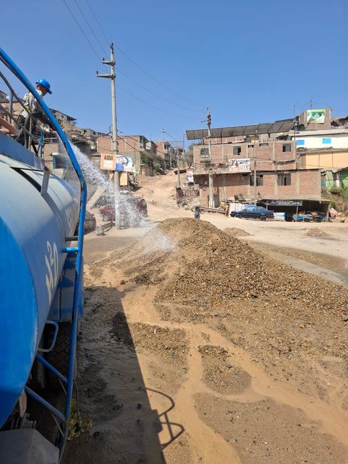 Un camión cisterna azul rocía agua sobre un camino embarrado en un barrio polvoriento de ladera bajo un cielo despejado.