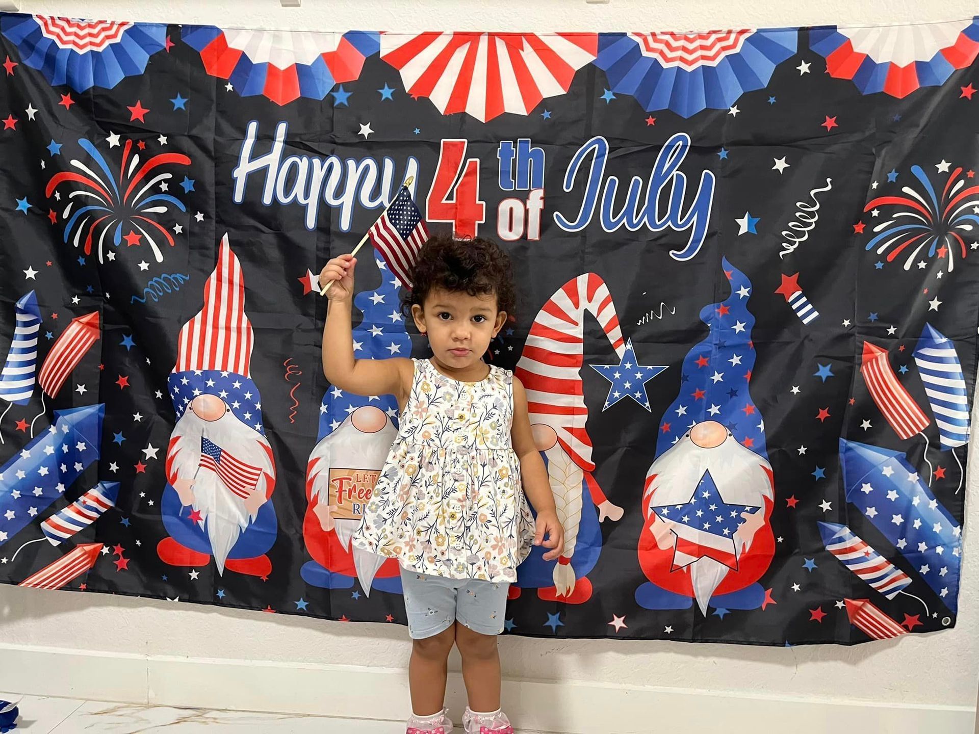 A little girl is standing in front of a happy 4th of july banner.