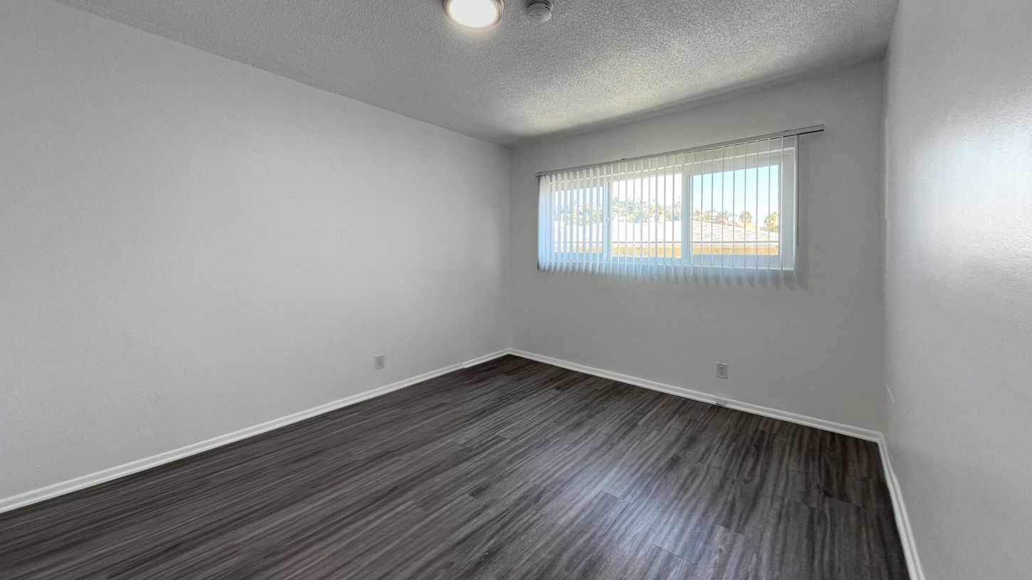 Empty bedroom with gray wood-look floor, white walls, and a window with sheer curtains.