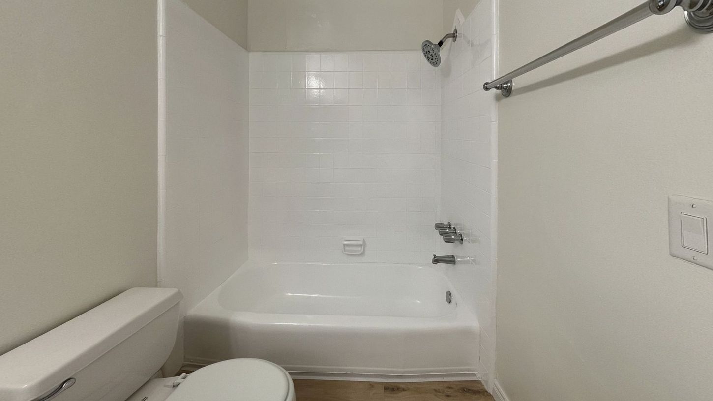 White bathroom with a tub, shower, toilet, towel rack, and a light switch on the wall.