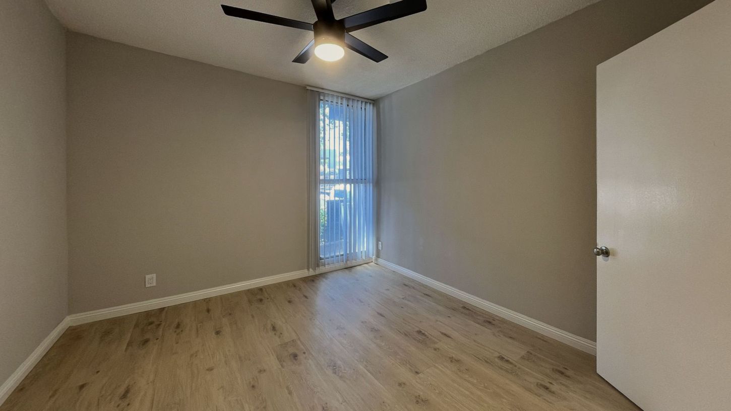 Empty bedroom with beige walls, wood flooring, a window with curtains, and a ceiling fan.
