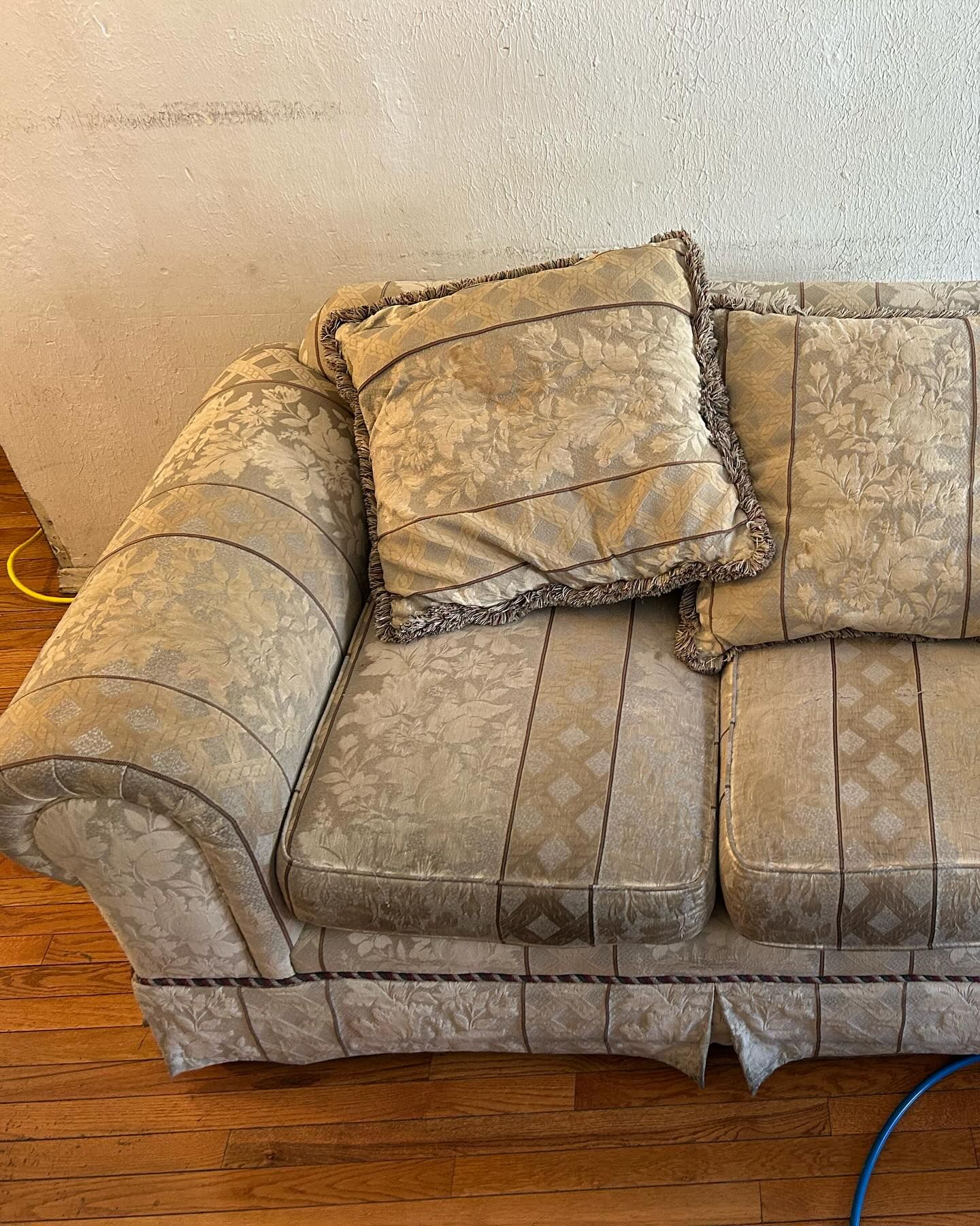 Worn beige couch with patterned cushions against a wall on a hardwood floor