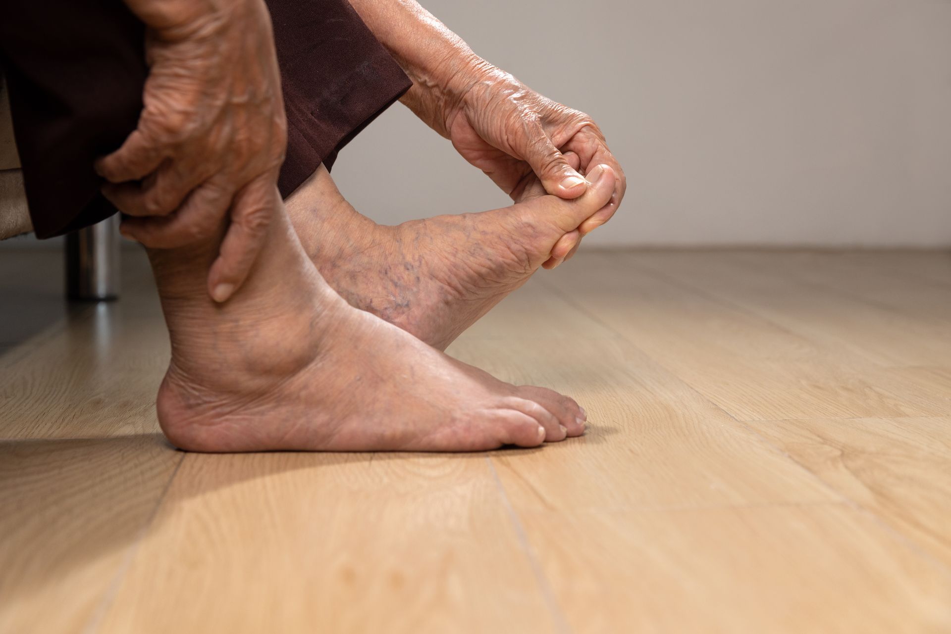 Person holding their swollen ankle and foot, sitting on a wooden floor, possibly experiencing pain.