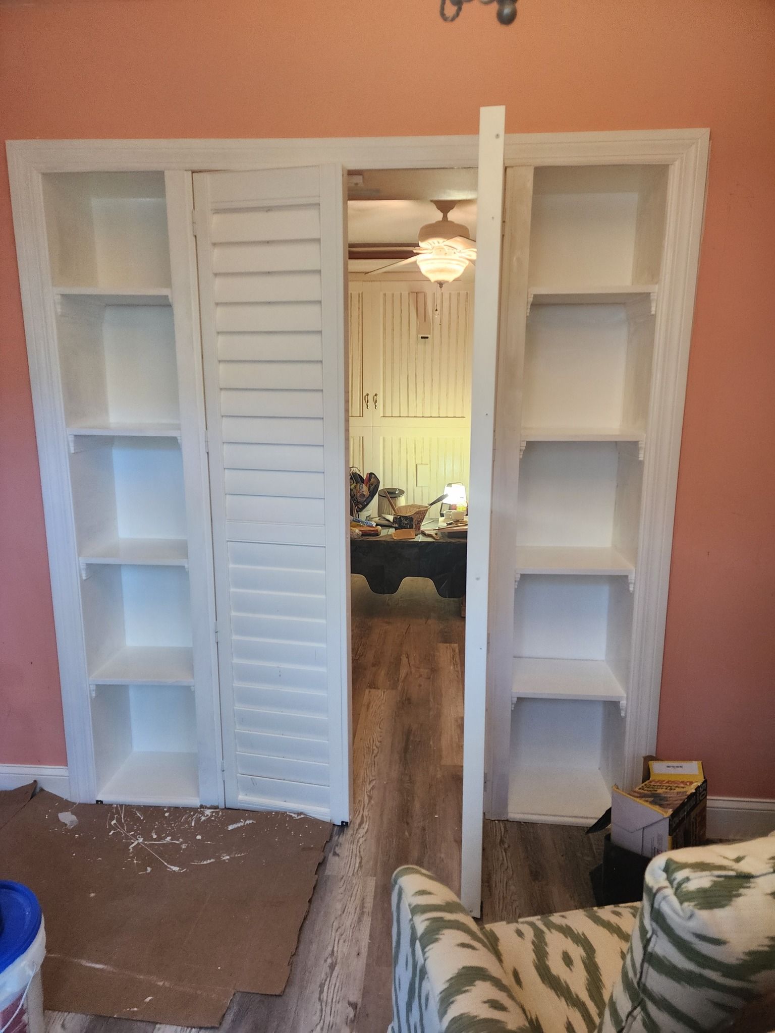 White built-in bookshelves frame a doorway. One shutter door is partially open, revealing a room with a table and chair.