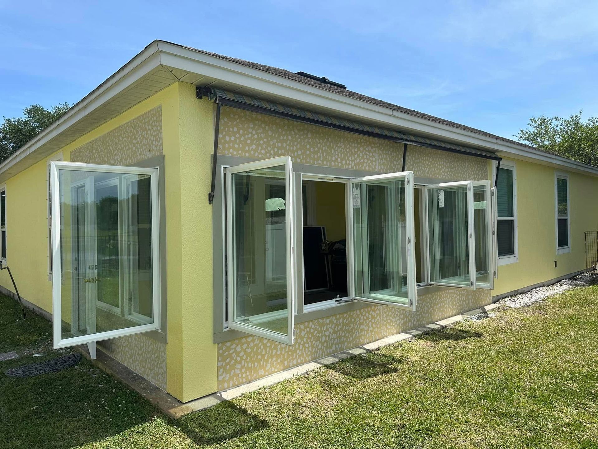 Yellow house exterior with several open white-framed windows, green grass, and blue sky.