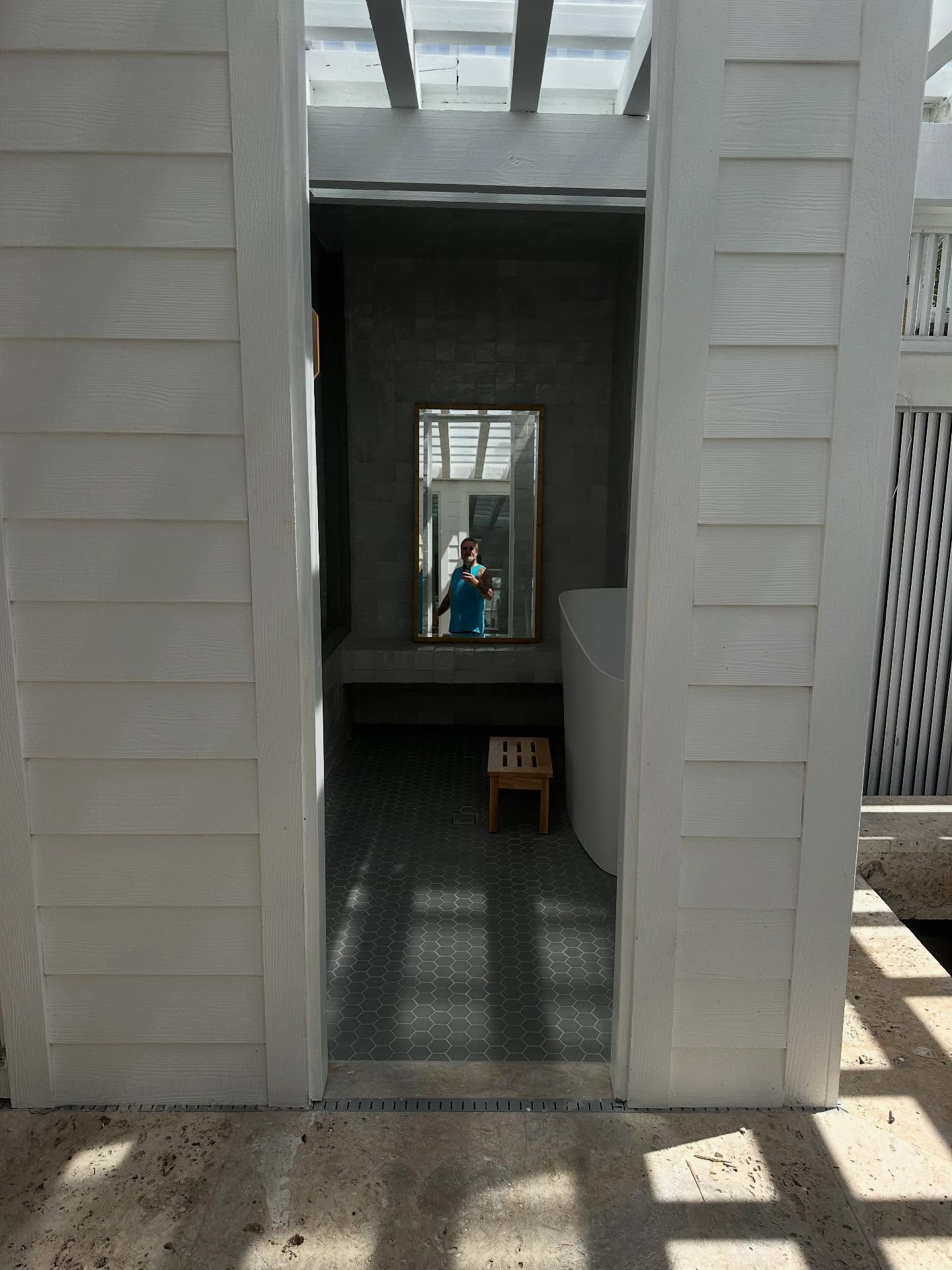 White-walled room entrance, mirror reflects person. Small wooden stool, light tile floor.