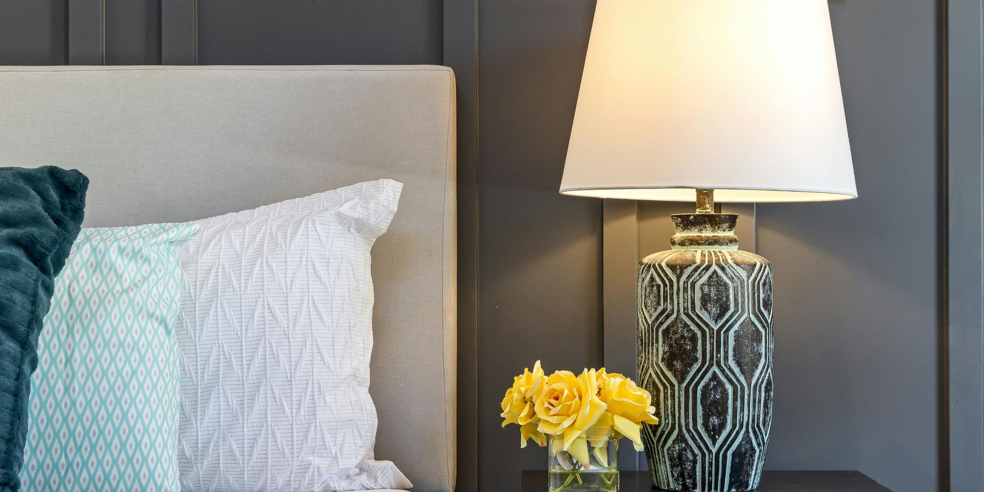 Bedroom nightstand with a lamp and flowers. Pillows rest against a headboard.
