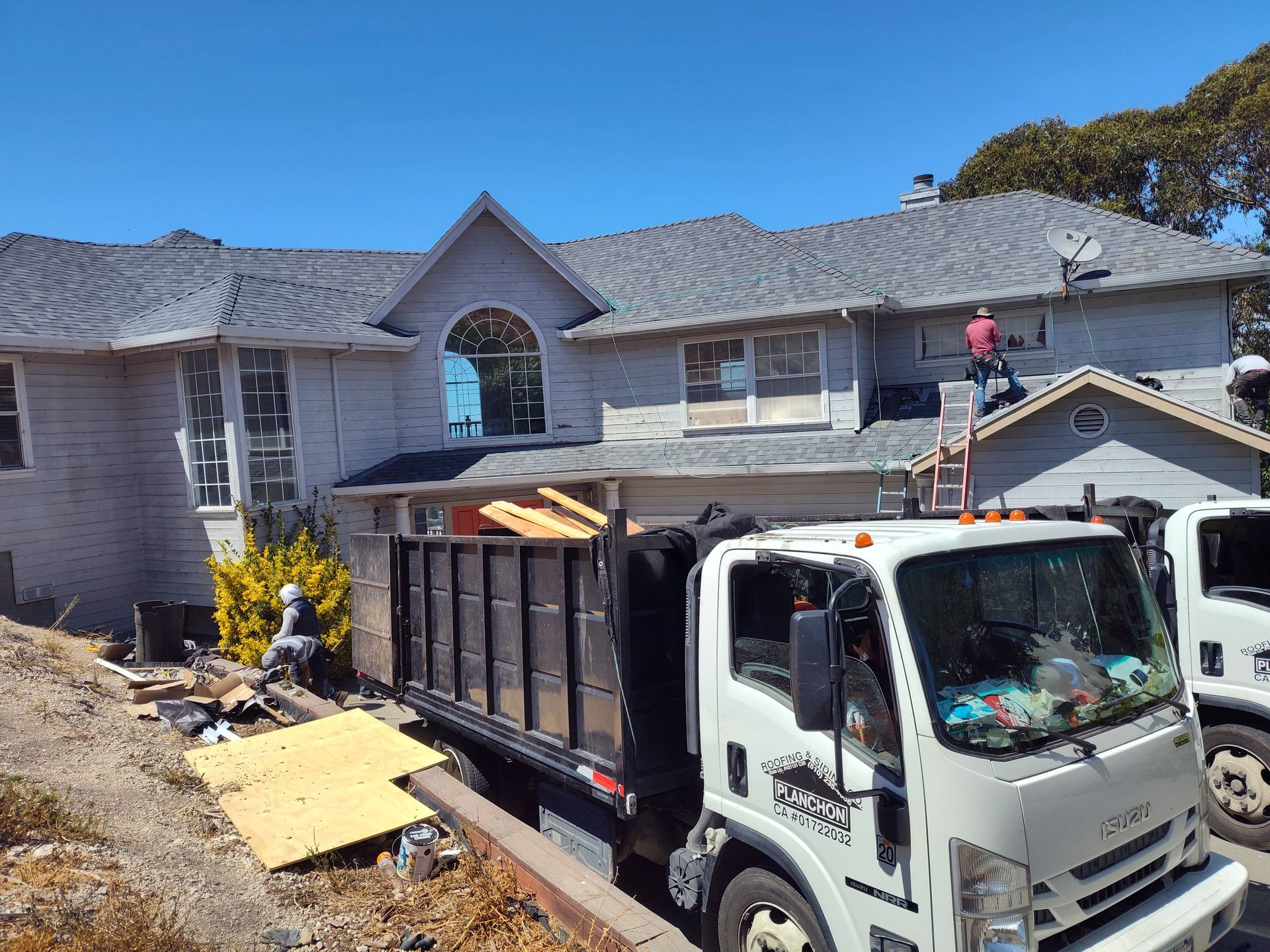 House with workers on the roof; dump truck in front; blue sky.