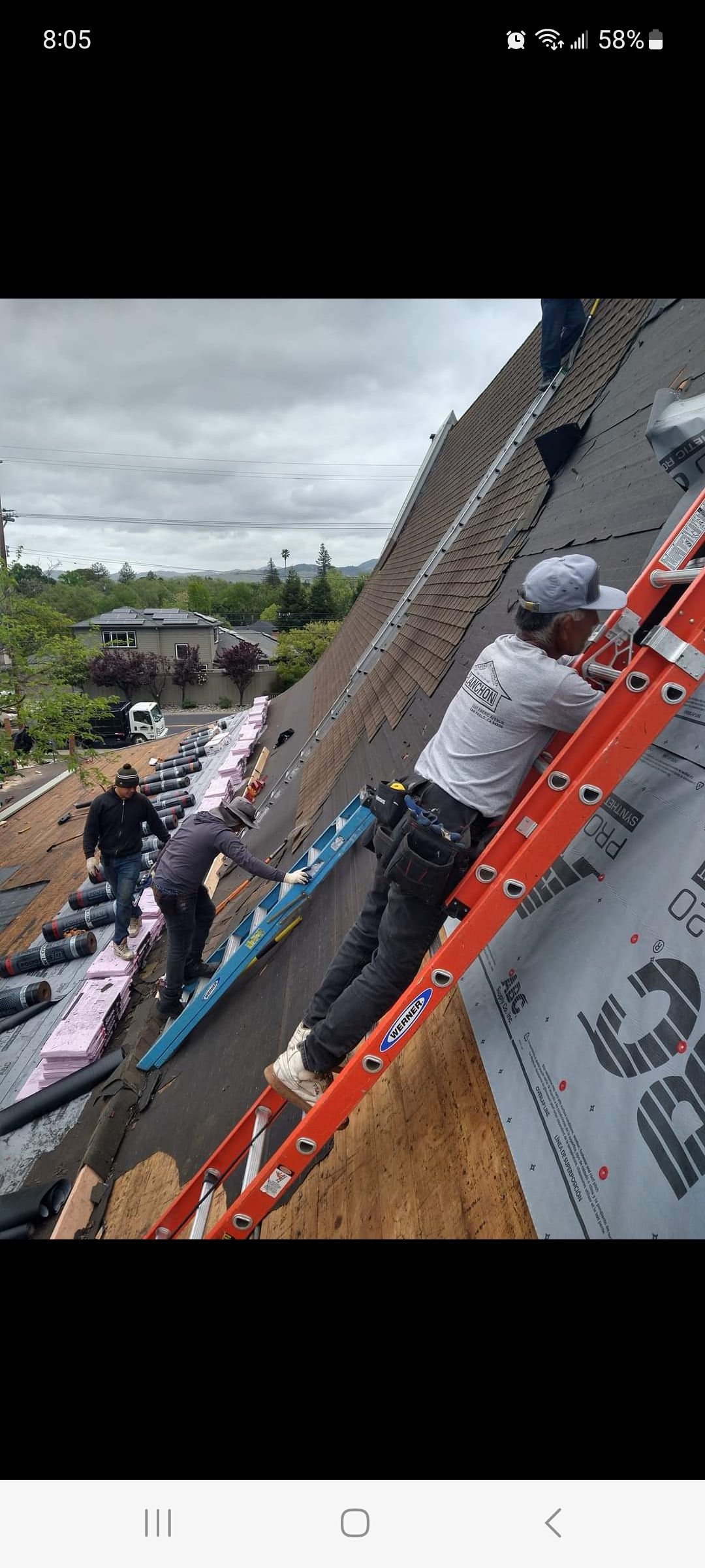 Roofers working on a steep roof, one on a ladder. Overcast day with a partial view of houses and trees.