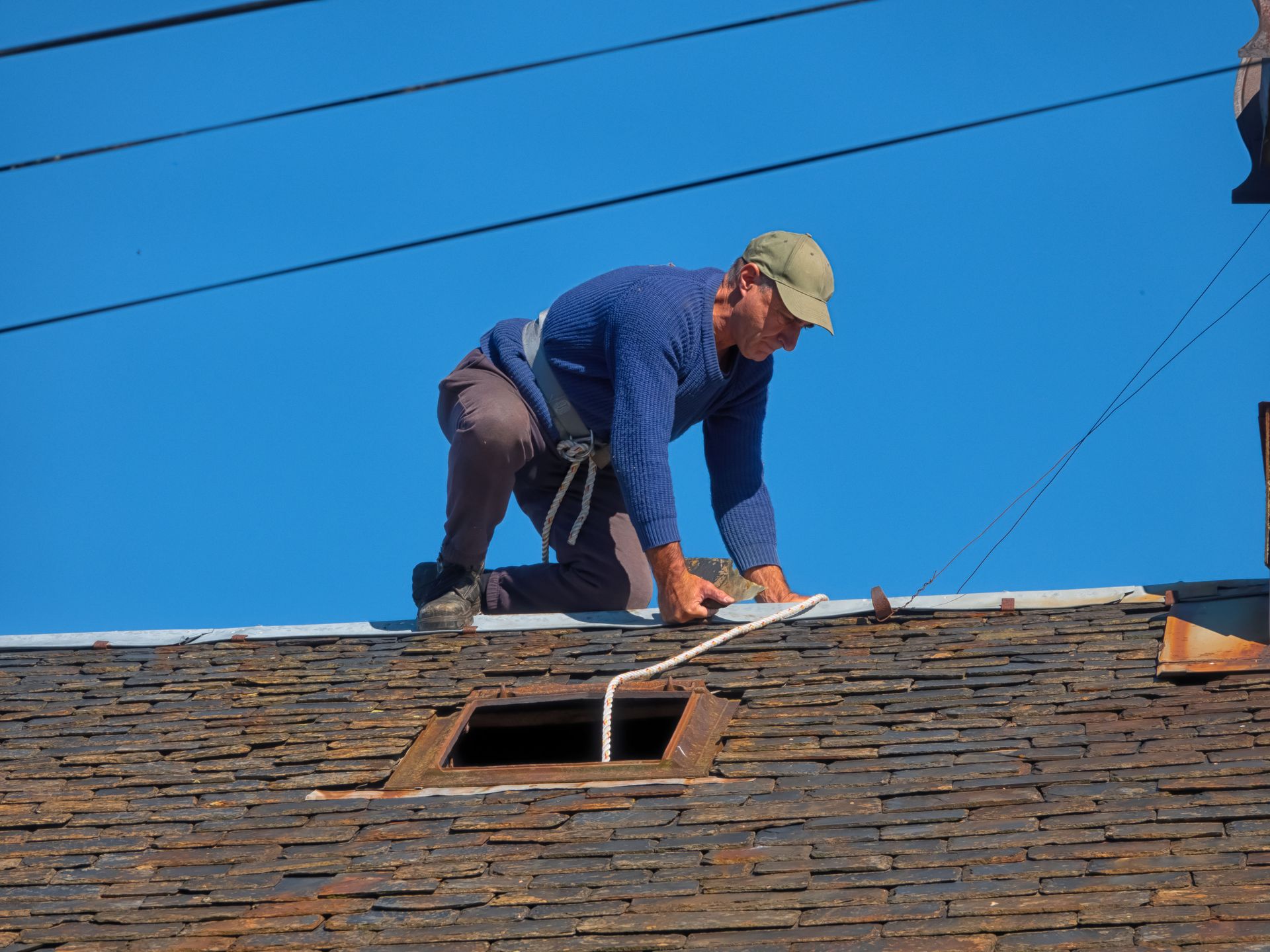 Roofing contractor securing safety rope while working on slate roof under clear blue sky.