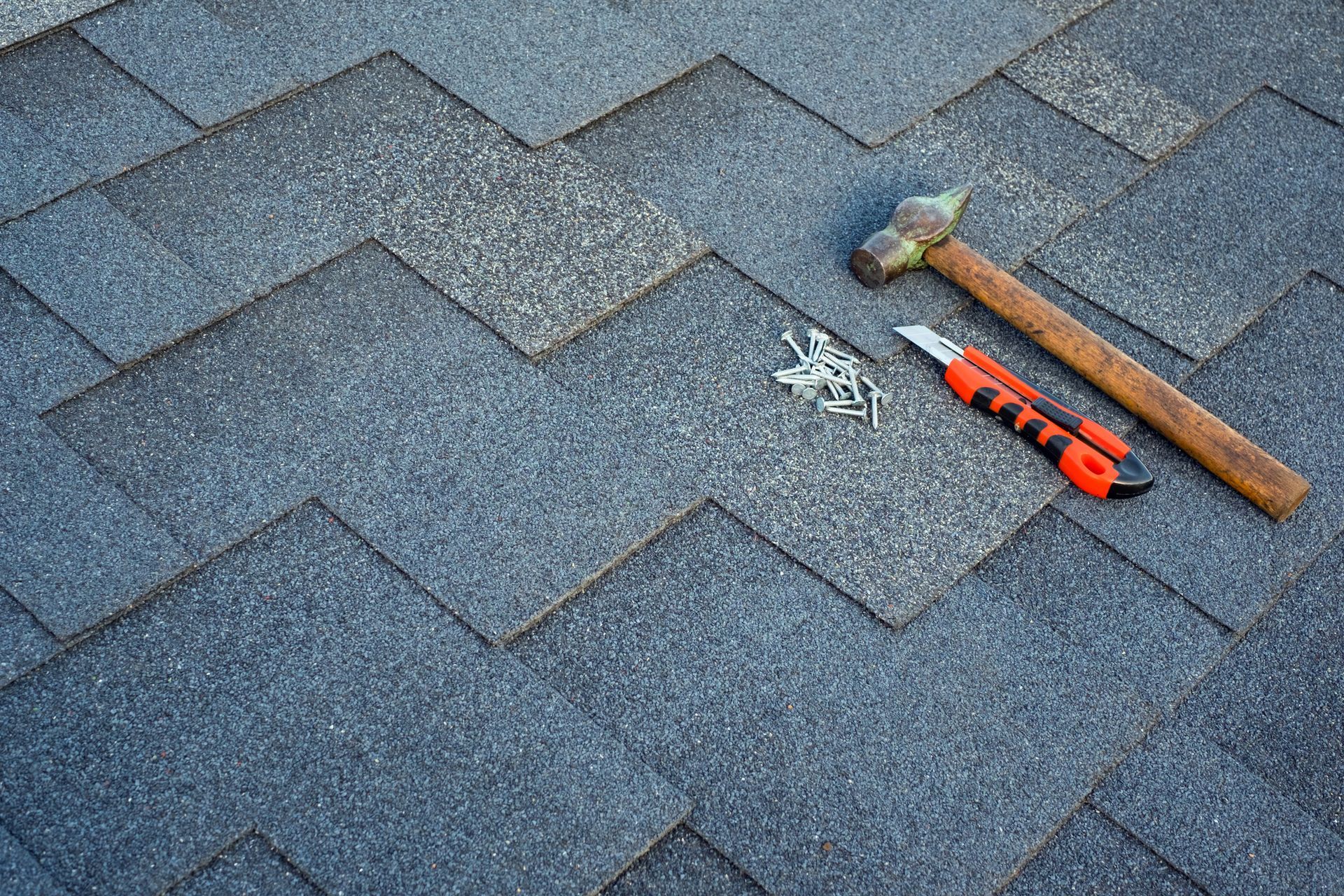 Roof shingles repair tools including hammer, utility knife, and nails on asphalt roofing.
