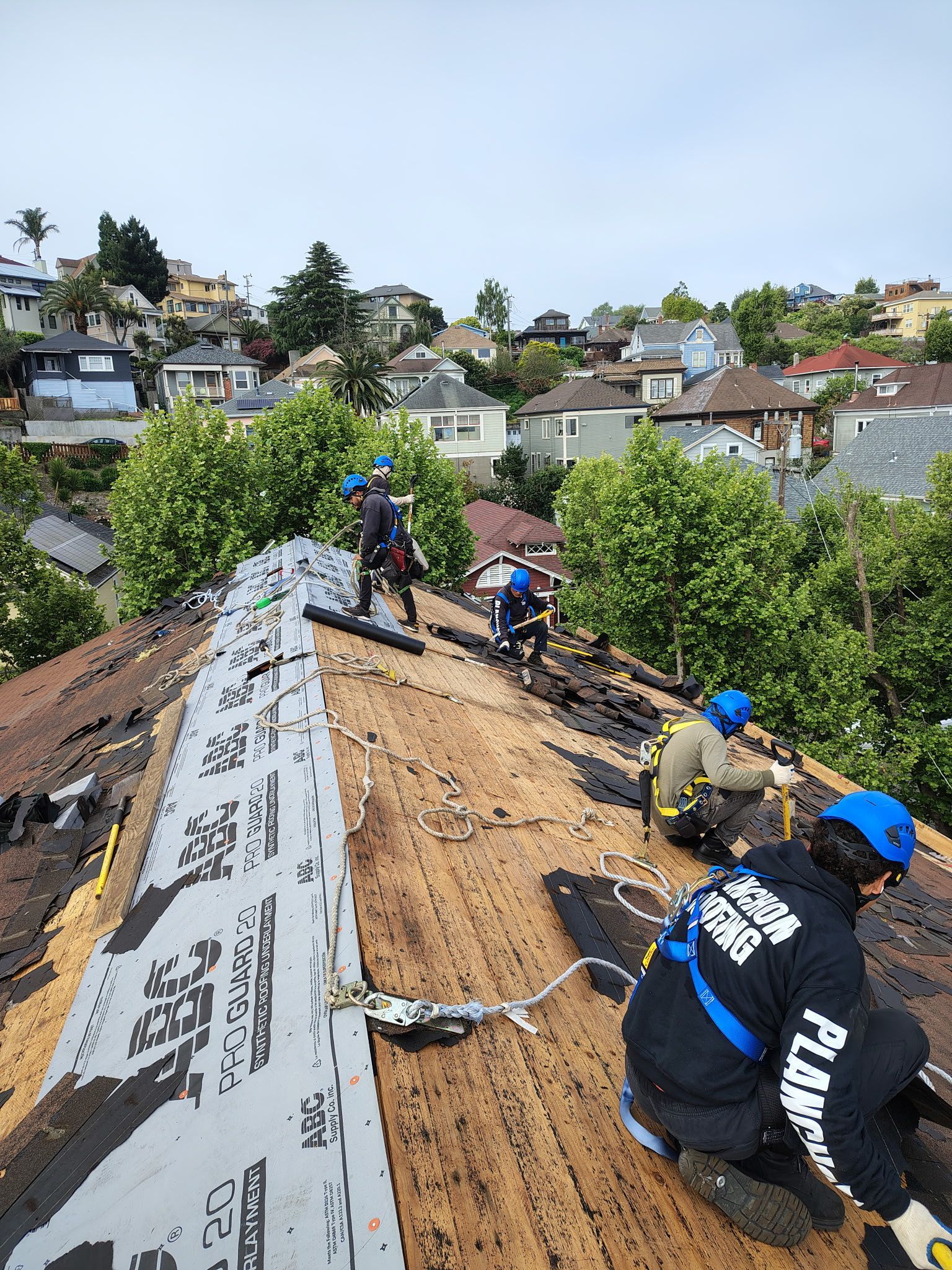 Roofers working on a residential roof, wearing safety harnesses. Houses and trees in the background.