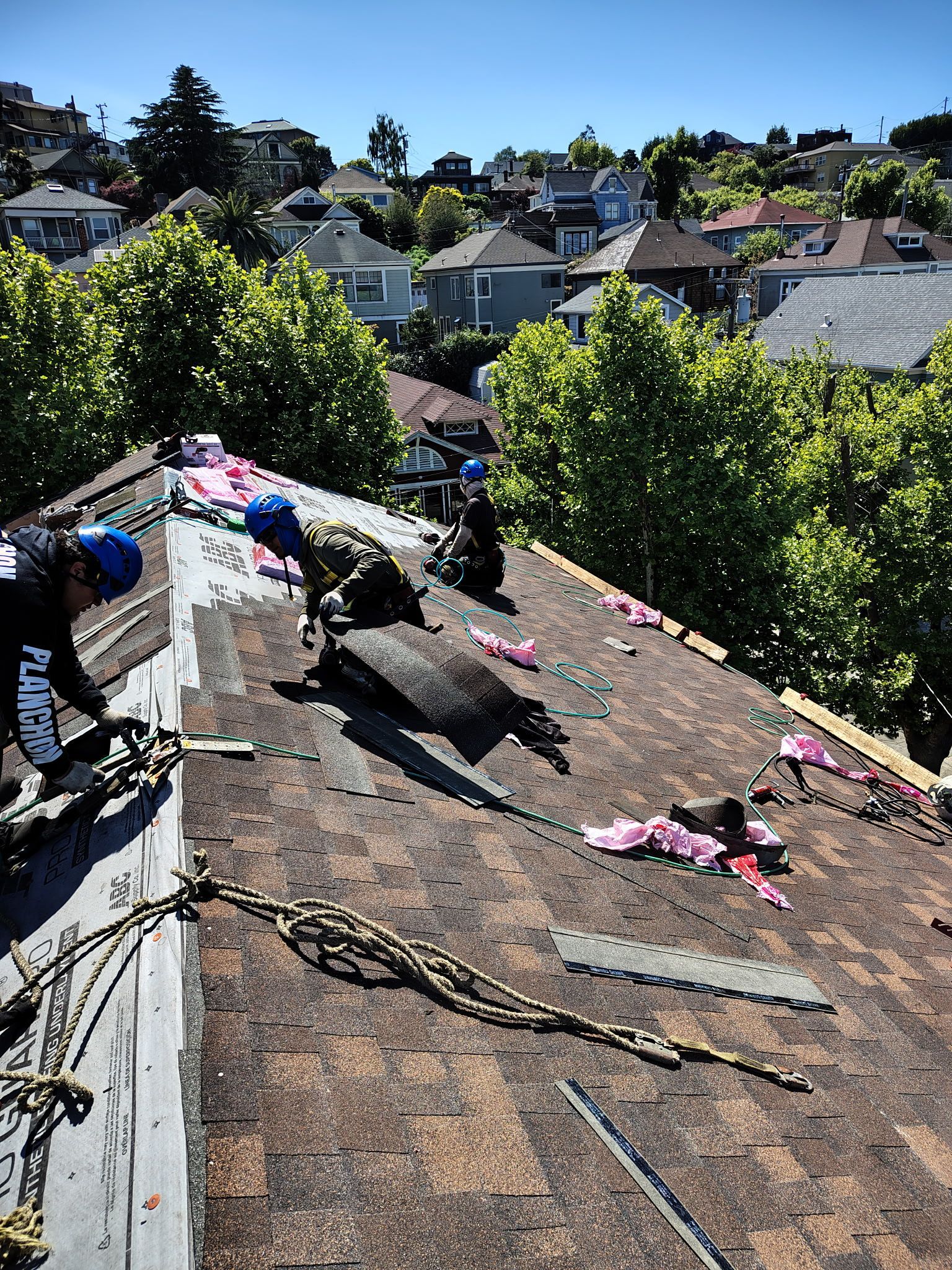 Roofers working on a residential roof, removing old shingles on a sunny day. Houses and trees in background.