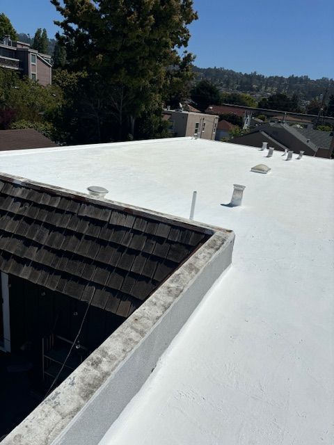 White flat roof on a building, with vent pipes. Dark wood shingle roof in the foreground. Green trees and hills in the background.
