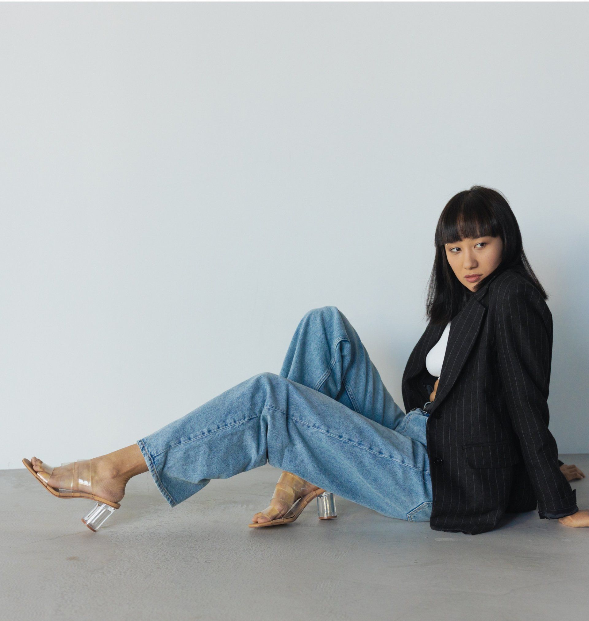Person wearing light blue jeans and heels with a black blazer, standing against a plain wall.