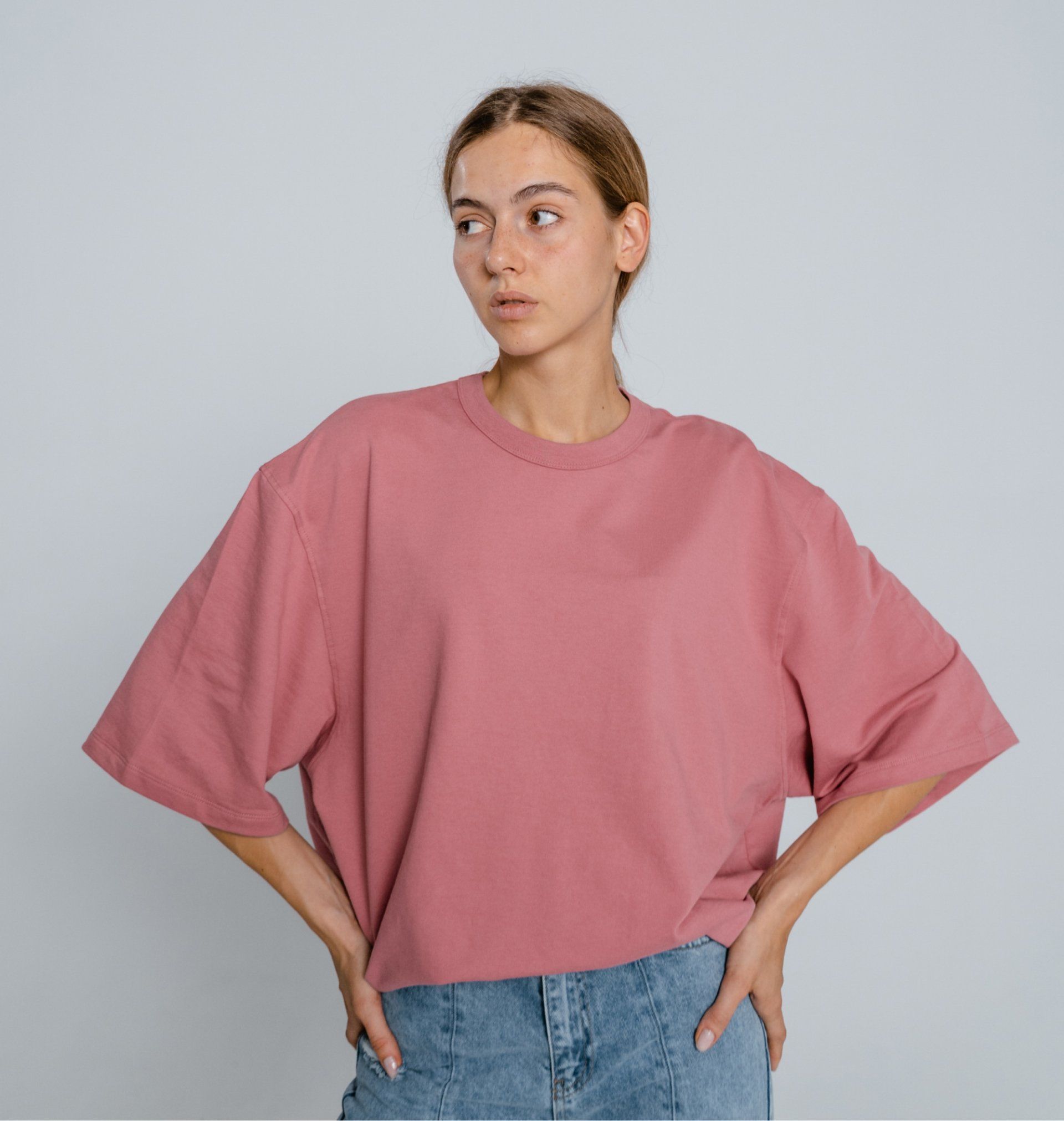Person in a pink oversized T-shirt and jeans standing with hands on hips, looking to the side.