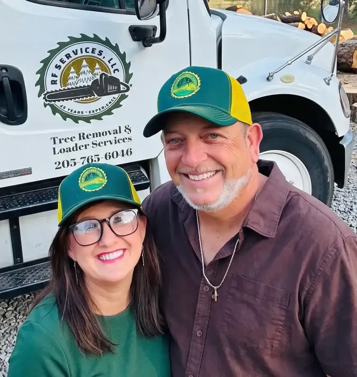 Smiling person in a green cap and yellow shirt standing outdoors by a tree and shrubs.