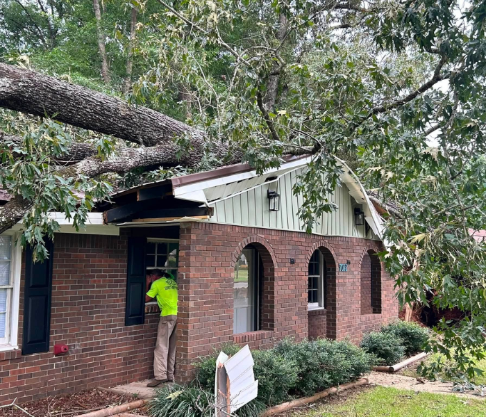 Worker in bright green shirt inspecting storm-damaged brick house with fallen tree and broken roof