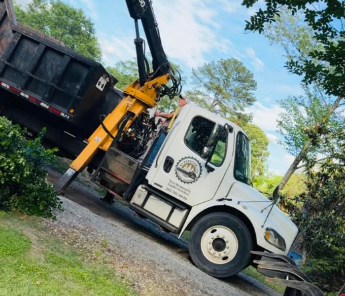 White dump truck with crane arm on a dirt road beside trees and grass, viewed at an angle