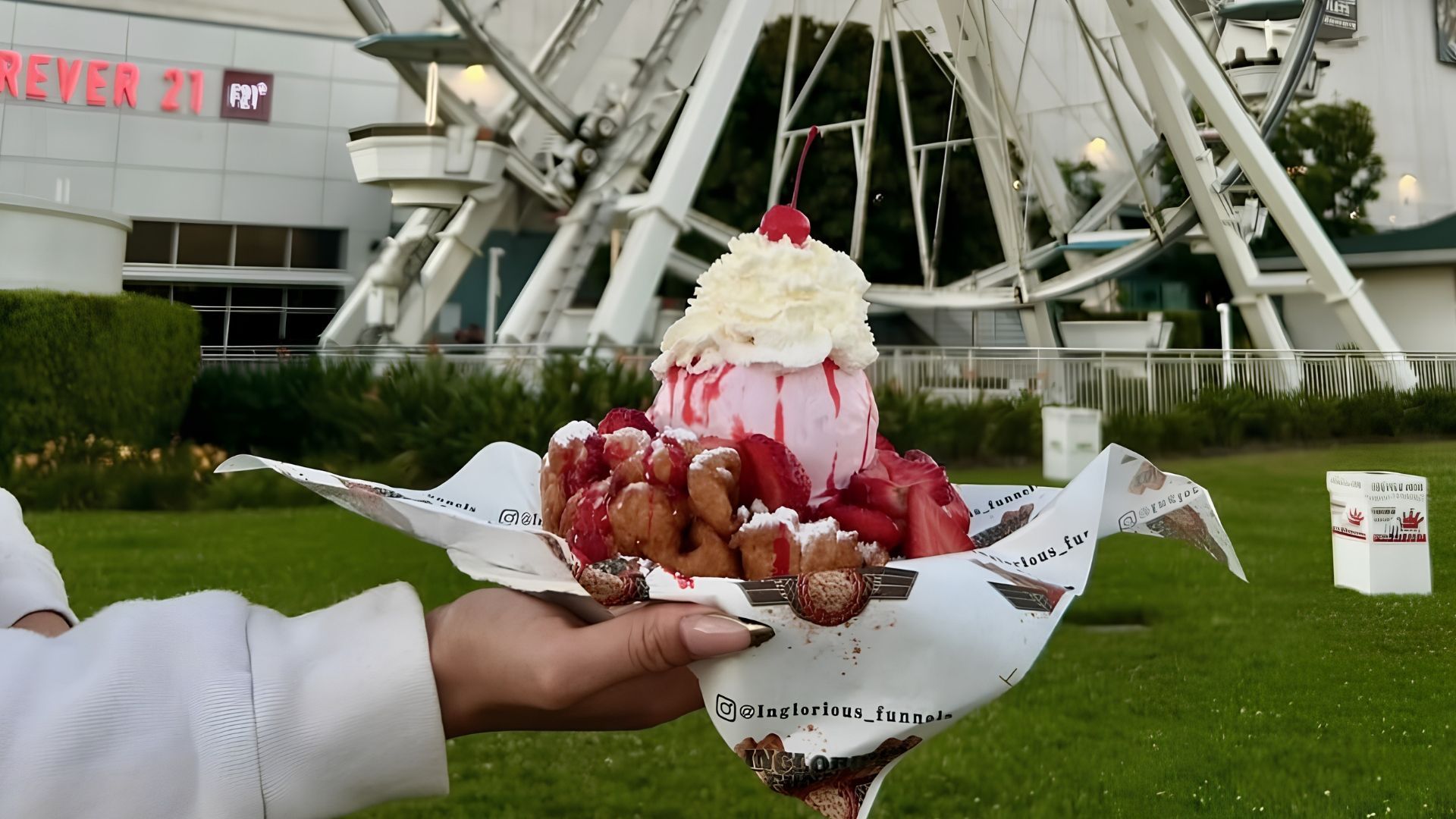 Hands holding a tray of strawberry-topped shaved ice in front of a ferris wheel on grass.