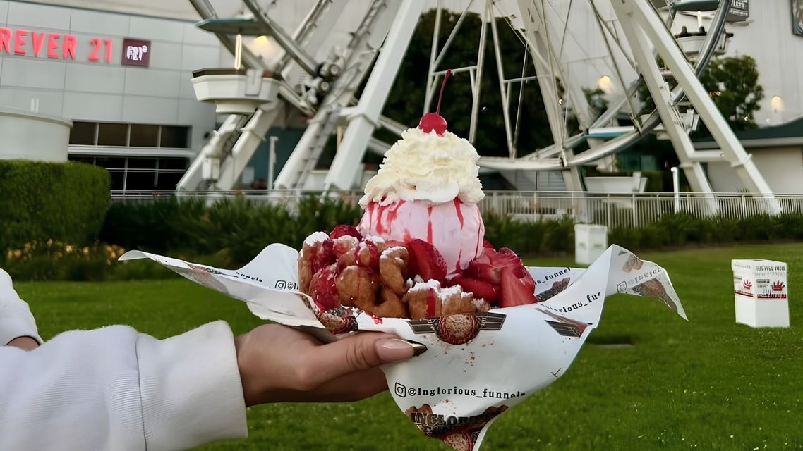 Hands holding a tray of strawberry-topped shaved ice in front of a ferris wheel on grass.