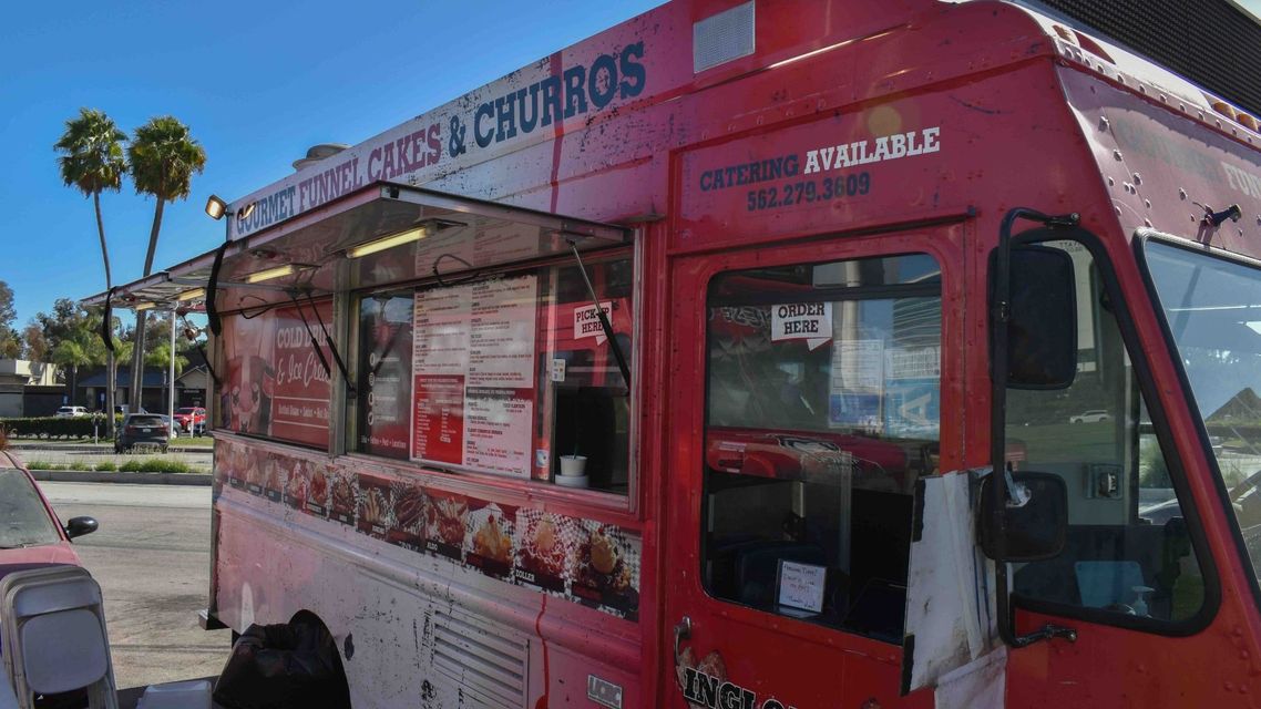 Red churros truck parked on a sunny street, with menu signs and a serving window open.