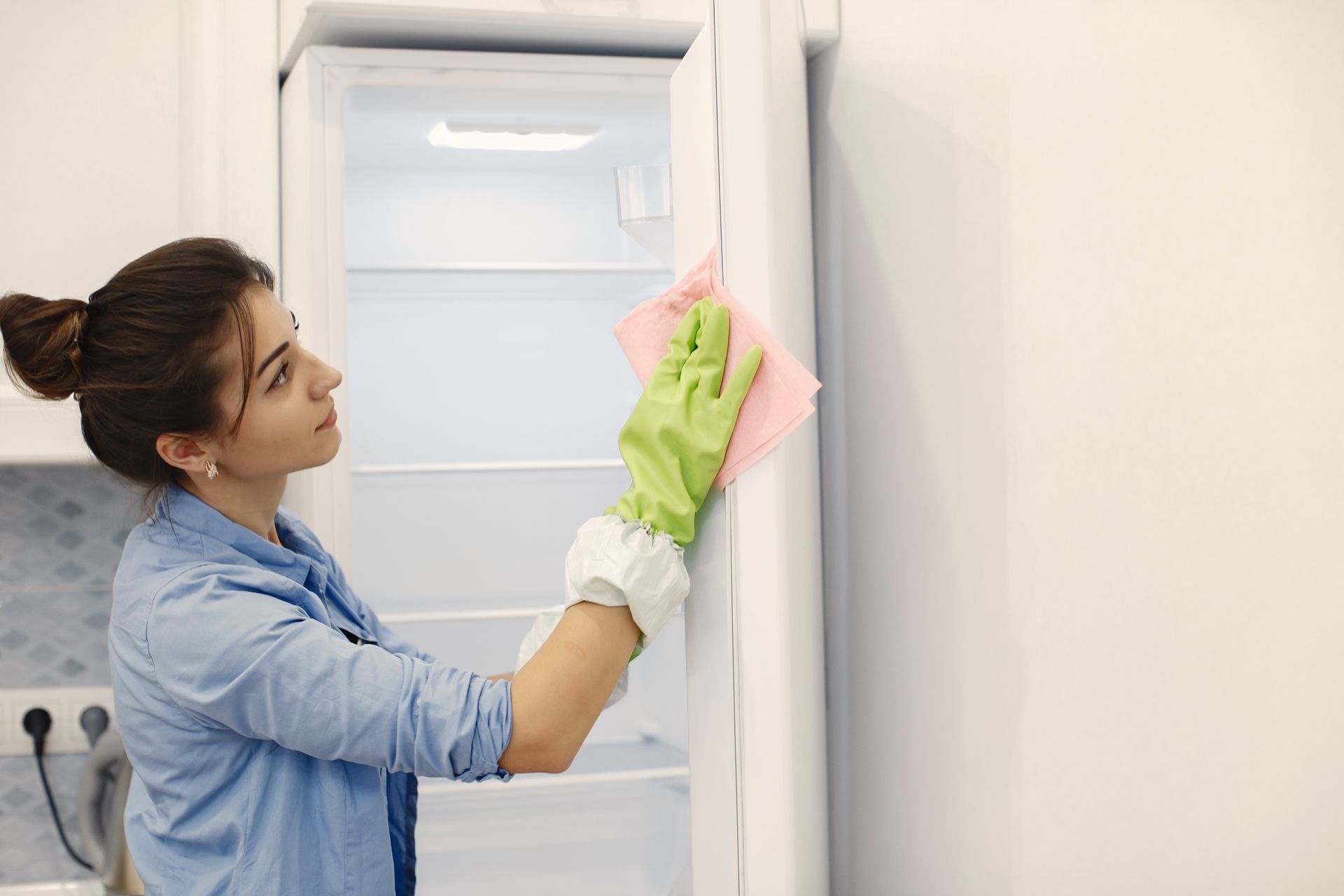 A woman is cleaning a refrigerator door with a sponge.