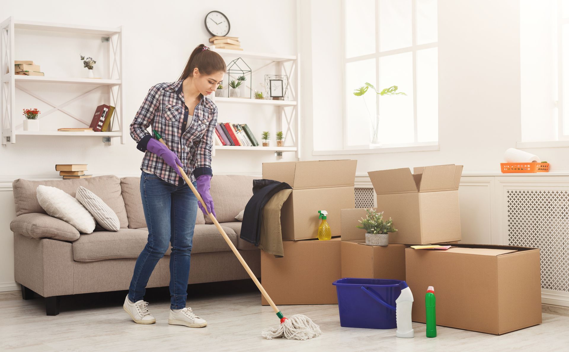 A woman is mopping the floor in a living room filled with boxes.
