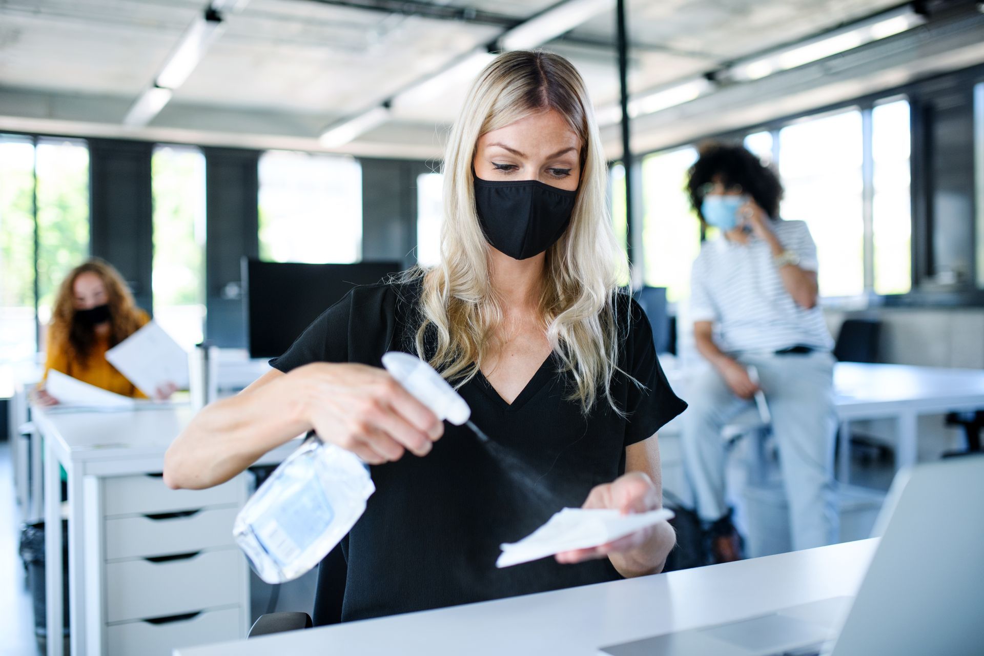 A woman wearing a mask is cleaning her desk in an office.