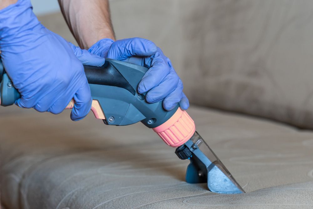 A Person Wearing Blue Gloves Is Cleaning A Couch With A Vacuum Cleaner — Big Red Carpet & Property Maintenance In Collinsville, QLD A Person Wearing Blue Gloves Is Cleaning A Couch With A Vacuum Cleaner — Big Red Carpet & Property Maintenance In Collinsville, QLD