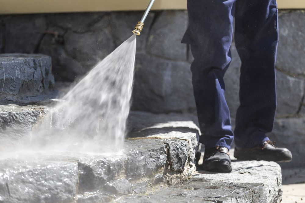 A Person Is Cleaning A Stone Wall With A High Pressure Washer — Big Red Carpet & Property Maintenance In Collinsville, QLD A Person Is Cleaning A Stone Wall With A High Pressure Washer — Big Red Carpet & Property Maintenance In Collinsville, QLD
