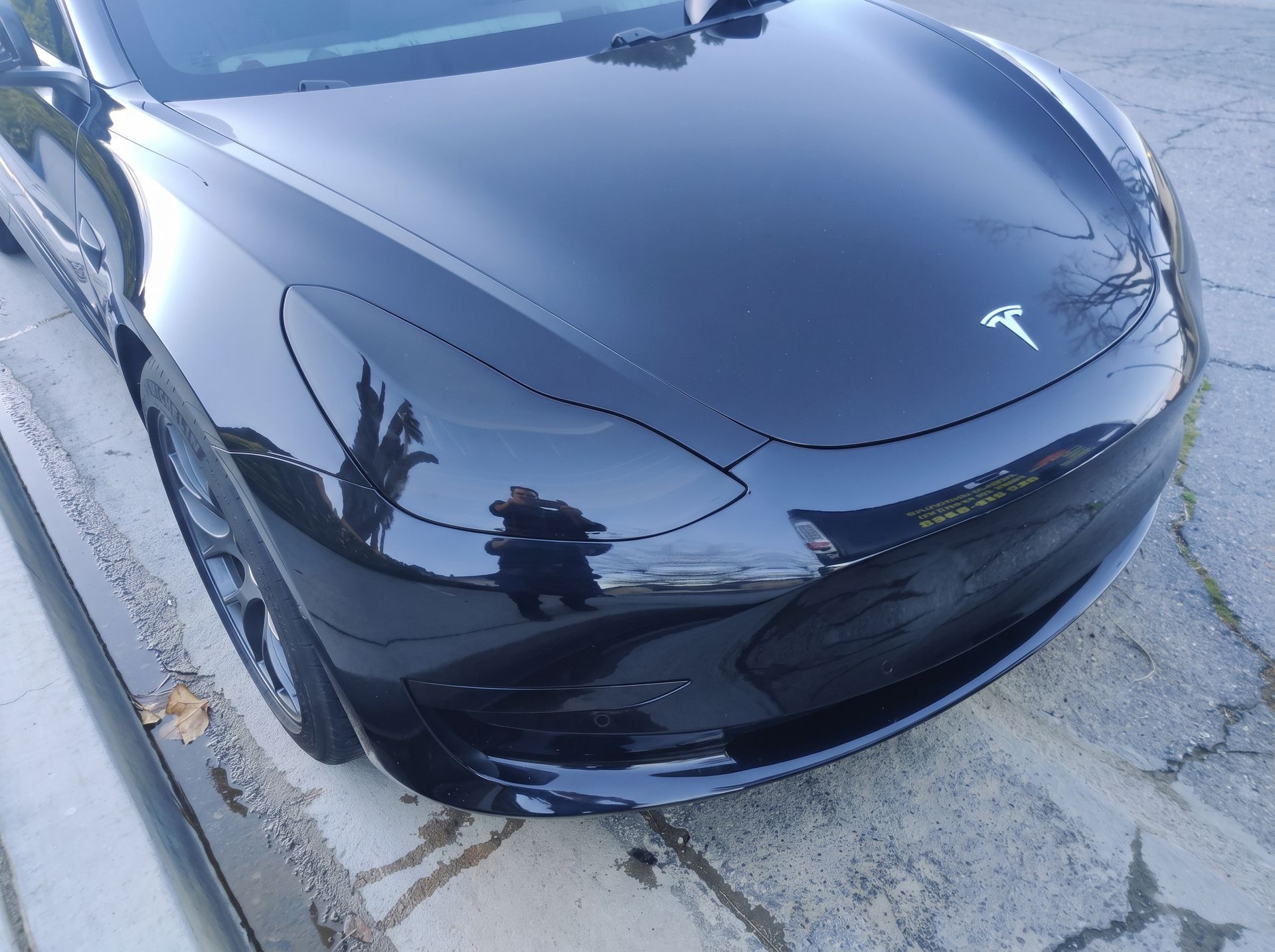 Black Tesla car viewed from the front, parked on a concrete driveway with visible reflections on the hood
