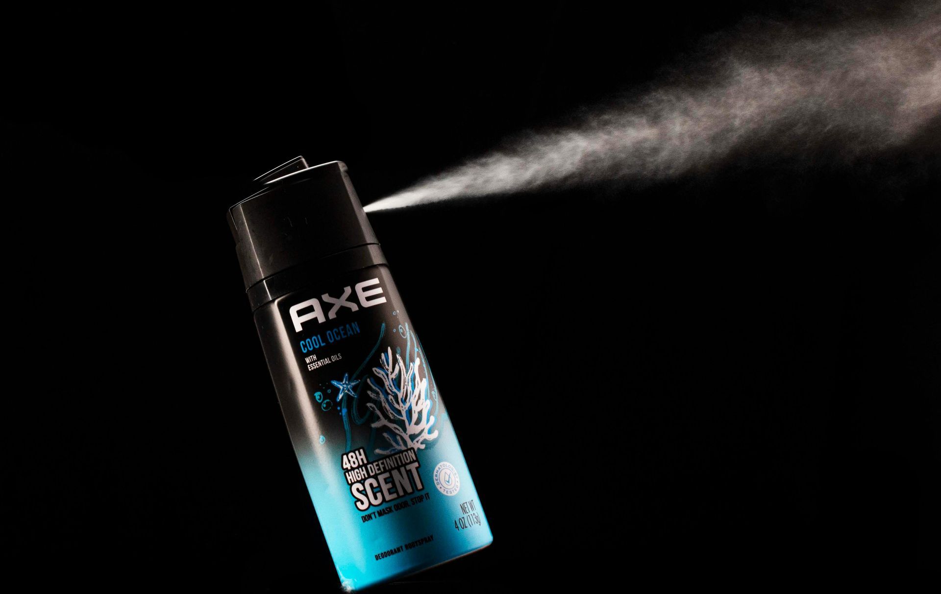 A black and teal Axe body spray aerosol  can angled to the left, spraying a mist into a dark background.