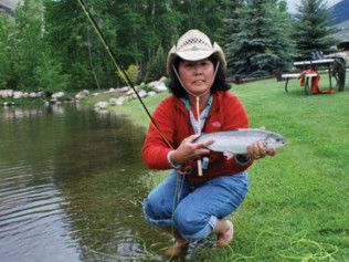 A woman in a cowboy hat is kneeling in the grass holding a fish.