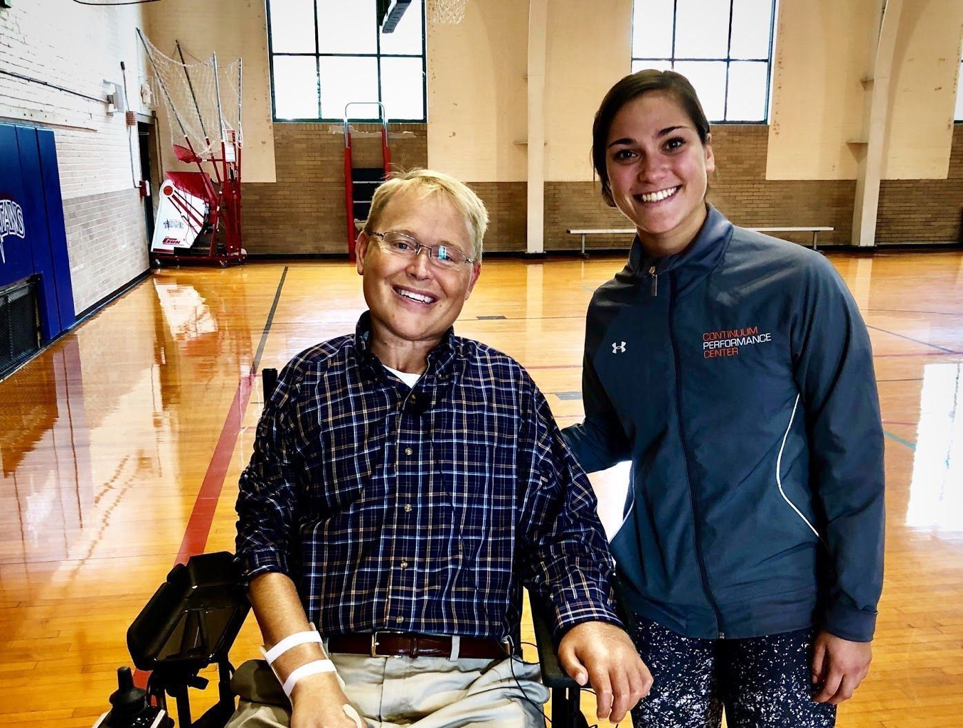 Two people smiling in a gym, one seated in a wheelchair, with basketball hoops in the background