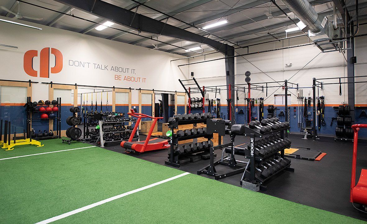 Spacious gym with turf, weight racks, benches, and bright orange equipment under high ceilings.