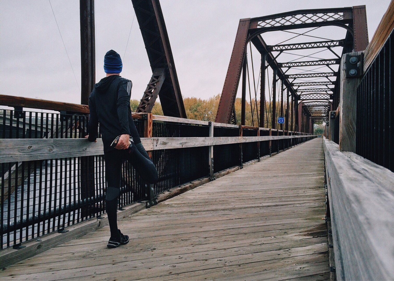 Person on a wooden bridge in a blue hat, holding the railing and walking toward the iron truss span.