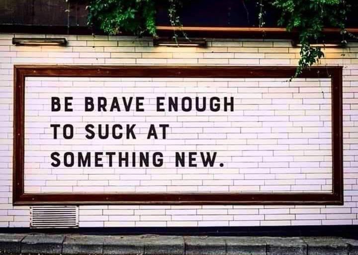 Wall sign with bold black text: “BE BRAVE ENOUGH TO SUCK AT SOMETHING NEW.”