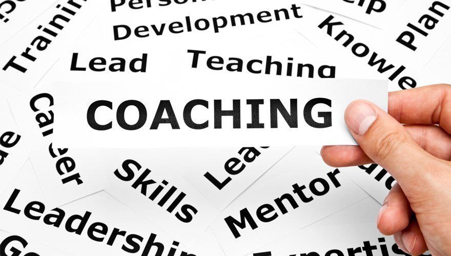 Hand holding a magnifying glass over the word “COACHING” surrounded by related business terms