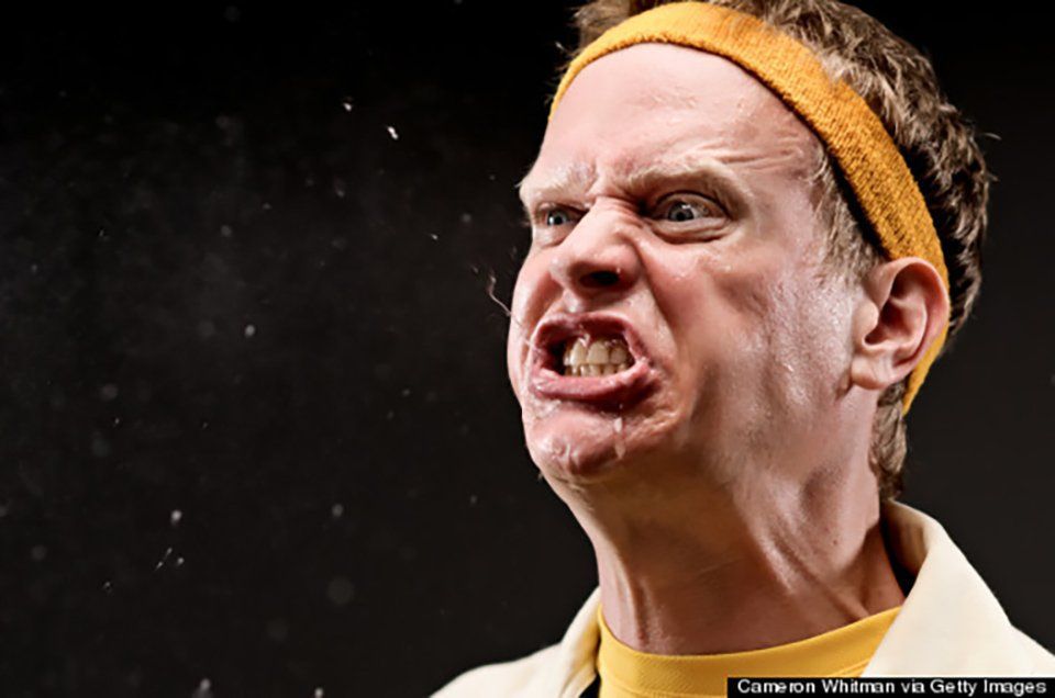 Close-up of a person grimacing on a dark stage, wearing a yellow headband and beige shirt